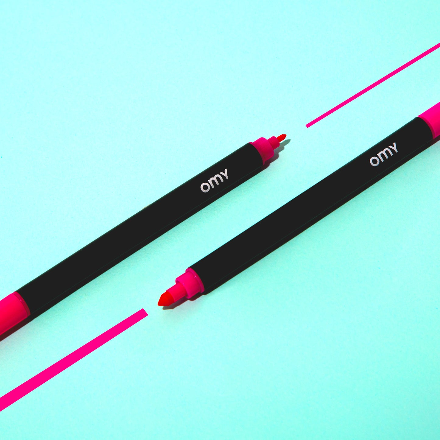 Two OMY black markers with pink tips on a light blue background, showing both tips.